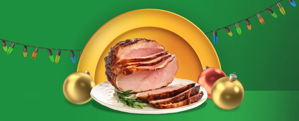 A joint of ham is sat on a serving plate. Christmas baubles and lights also feature. On a two-tone green background.