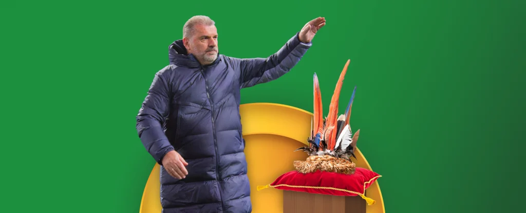 Ange Postecoglou, the coach of the Tottenham Hotspur football team wearing a dark blue jacket and raising his hand. Beside him is a wooden podium with a red cushion, topped with a crown like arrangement of colorful feathers on a vibrant green background with a yellow semi circle framing the feather arrangement.